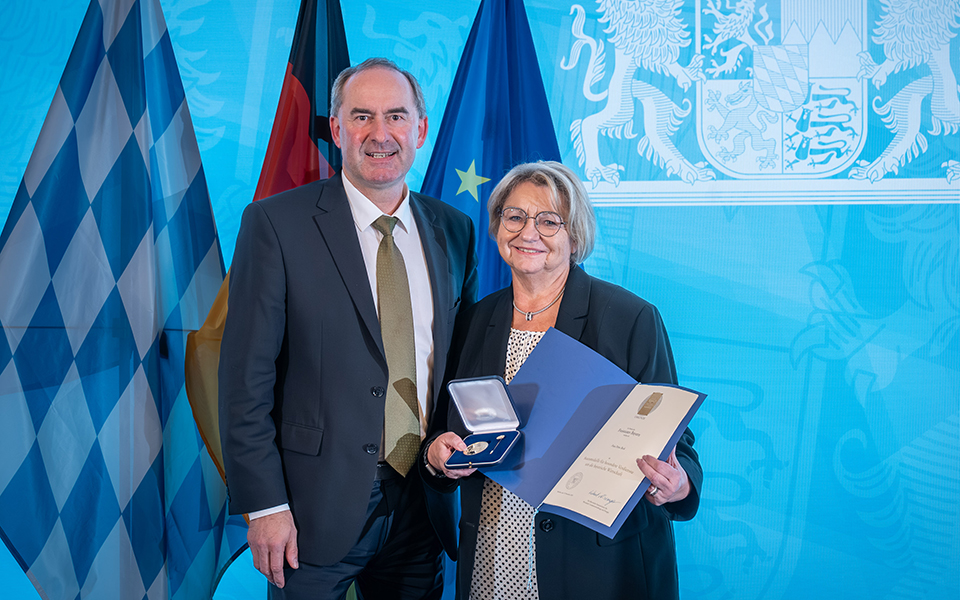 Petra Beck mit Staatsmedaille und Urkunde in den Händen, daneben Staatsminister Hubert Aiwanger, im Hintergrund die bayerische, die deutsche und die Europaflagge sowie das Wappen des Freistaats Bayern