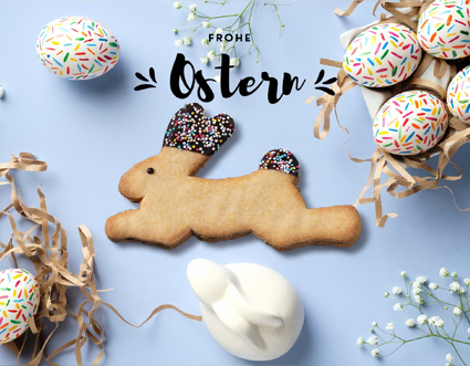 Ein Hasi-Keks liegt zwischen bemalten Ostereiern und Osterdeko auf blauem Untergrund