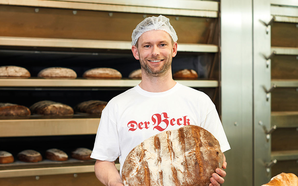 Bäcker hält ein Brot in der Hand, im Hintergrund  Backofen gefüllt mit Broten